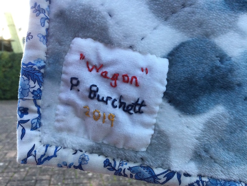 May include: A white fabric label with blue, red, and gold embroidery. The label reads "Wagon" P. Burchett 2019.