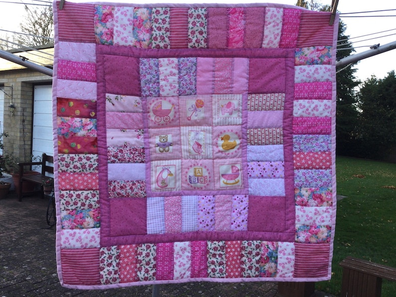 Op de afbeelding: Een roze en witte patchwork babydeken met verschillende bloemenpatronen en geborduurde afbeeldingen van een kinderwagen, een teddybeer, een eend, een schommelpaard en blokken die "A B C" spellen.