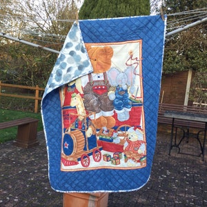 May include: A blue and white quilted baby blanket featuring a colourful print of a bear, elephant, and other animals playing with toys. The blanket has a blue and white polka dot pattern on the reverse side.