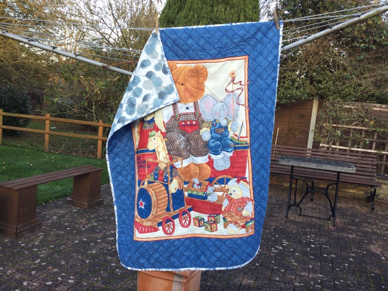 May include: A blue and white quilted baby blanket featuring a colourful print of a bear, elephant, and other animals playing with toys. The blanket has a blue and white polka dot pattern on the reverse side.