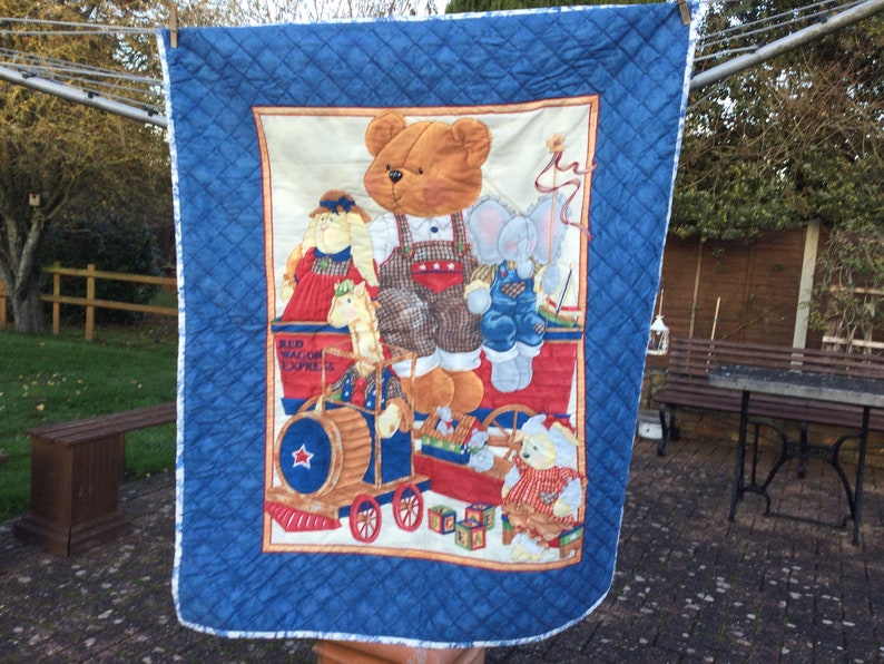 May include: A blue and white quilted baby blanket with a colourful print of stuffed animals riding in a red wagon. The wagon says "Red Wagon Express".