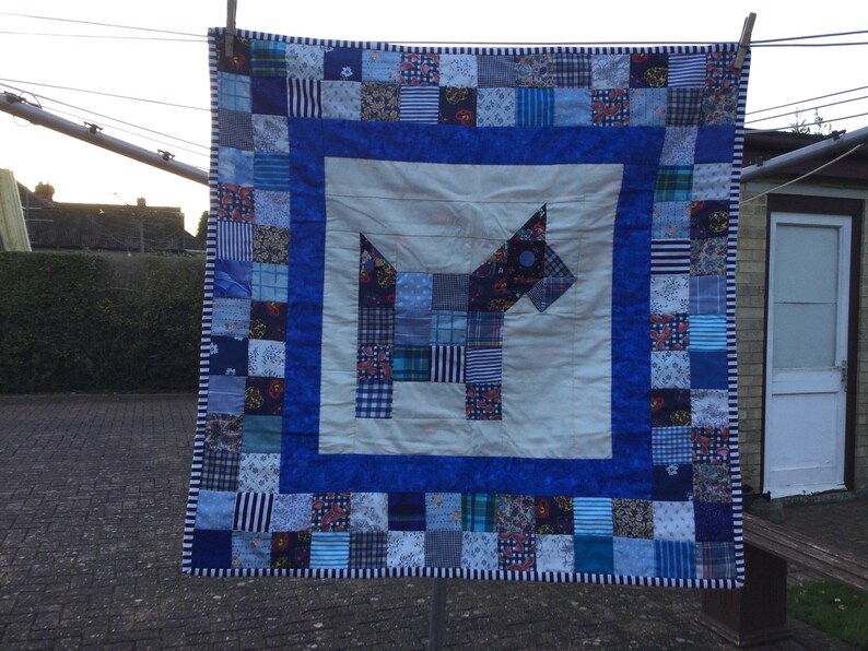 K&ouml;nnte beinhalten: Eine blaue und wei&szlig;e Patchwork-Decke mit einem blauen Hundedesign in der Mitte. Die Decke besteht aus Quadraten aus verschiedenen Stoffen und ist von einem blauen Rand umgeben.