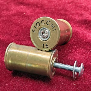 Puede incluir: Dos casquillos de escopeta de latón reutilizados como herrajes. El casquillo superior está marcado como Fiocchi 16 con un centro plateado. El casquillo inferior presenta un perno de metal plateado roscado con arandela y tuerca a juego.