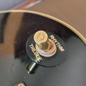 May include: Close-up of a black electric guitar with a gold-colored bullet-shaped switch. The switch is labeled "Treble" and "Rhythm" in gold lettering. The guitar's body is glossy black.
