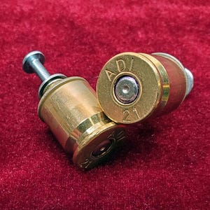 May include: Two brass-colored metal bullet casings repurposed as hardware, each featuring a silver-toned screw protruding from the center. The base of the casings displays the stamped text ADI and 21, with a metallic primer visible in the middle.