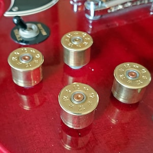 Puede incluir: Primer plano de una guitarra eléctrica roja con perillas personalizadas doradas con forma de cartuchos de escopeta. Las perillas son cilíndricas con un acabado de latón y presentan el número "12" en la parte superior. También se ve el interruptor y los herrajes cromados de la guitarra.