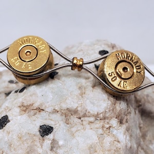 May include: A handcrafted bracelet with two brass bullet casings, each marked "HORNADY 50 AE." The casings are linked by silver wire and tiny gold beads. The bracelet is displayed on a textured, pale stone. The bracelet's design is unique.