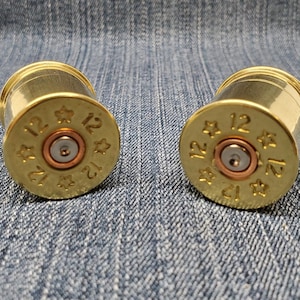 May include: Two brass-colored, cylindrical objects with a 12-gauge marking. The objects have a copper-colored center and are decorated with star-shaped imprints. The objects are placed on a blue denim surface.