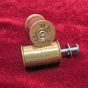 May include: Two brass-colored shotgun shell casings repurposed as hardware, each featuring a metallic base stamped with the text 20 GA FEDERAL. One casing is positioned vertically, while the other lies horizontally with a silver metal bolt and washer.