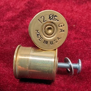 May include: Two brass shotgun shell-shaped cabinet knobs. The top knob is stamped with "12 GA" and "MADE IN U.S.A." The second knob has a screw and washer attached. The knobs are on a red velvet surface.