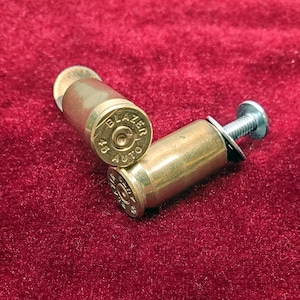 May include: Two brass-colored metal bullet casings with the text BLAZER 45 AUTO stamped on the base. One casing is fitted with a silver-colored metal screw and a flat washer protruding from the open end, creating a functional hardware component.