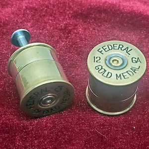 Puede incluir: Dos tapones de botella de latón con forma de cartuchos de escopeta. Uno tiene una tapa de rosca plateada. Ambos tienen el texto "FEDERAL 12 GA GOLD MEDAL" impreso. Sobre una superficie de terciopelo rojo.