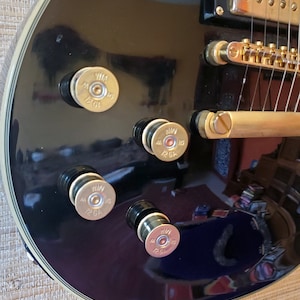 Peut inclure: Gros plan sur une guitare électrique noire avec des accastillages dorés. La guitare a quatre boutons dorés avec le texte "Win 12 GA" sur chaque bouton.