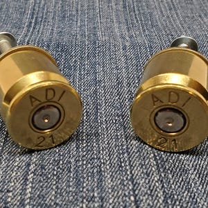 May include: Two brass-colored cabinet knobs with a bullet casing design. Each knob has the letters "AD/" and the number "21" stamped on the front. The knobs have a silver screw attached.
