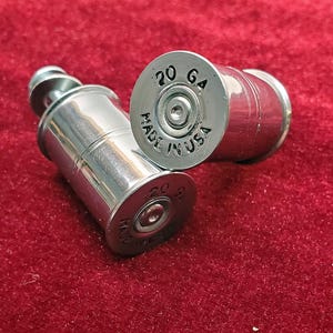 May include: Two polished silver-colored metal shotgun shell casings. The base of each casing is stamped with the text 20 GA and MADE IN USA. The cylindrical objects feature a smooth, reflective finish and a central primer indentation on the base.