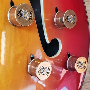 May include: Close-up of a guitar body with four custom knobs made from 12 GA shotgun shells. The guitar has a gradient finish, transitioning from orange to red. Each shell knob features a logo and the text "12 GA".