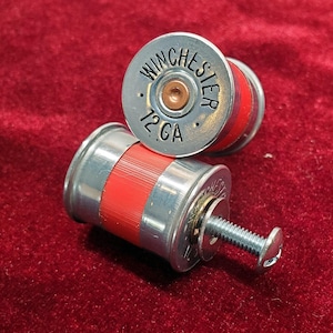 Puede incluir: Dos casquillos de escopeta Winchester 12 GA reutilizados y convertidos en herrajes. Cada casquillo presenta una base de metal plateado con el texto negro WINCHESTER 12 GA y un cuerpo de plástico rojo. Uno de los casquillos incluye un tornillo plateado.