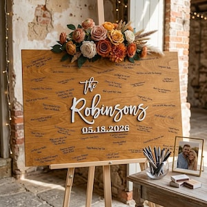 Op de afbeelding: Een rechthoekig houten gastenboekbord op een houten ezel, bekroond met een bloemstuk van oranje, witte en roze rozen. Het bord bevat de witte 3D-tekst The Robinsons en 05.18.2026, omringd door handgeschreven berichten in zwarte inkt.