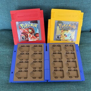 May include: Two Game Boy cartridges, one red and one yellow, featuring Pokémon game labels. A blue and brown cartridge holder is open, revealing slots for game storage. The red cartridge displays a Charizard, while the yellow one shows Pikachu.