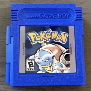 May include: A blue Nintendo Game Boy cartridge featuring the Pokémon game. The label shows the Pokémon logo and a blue turtle-like character. The cartridge has the text "Nintendo GAME BOY" and "THIS SIDE OUT". The game is rated "EVERYONE" by the ESRB.