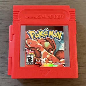 May include: A red Game Boy cartridge featuring the Pokémon game. The label displays the Pokémon logo and a Charizard illustration. The cartridge has the Nintendo Game Boy logo and the ESRB rating.