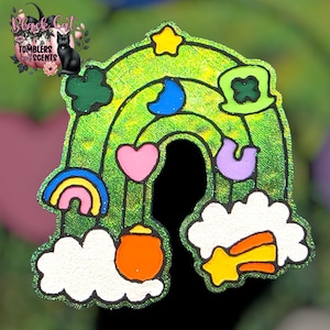 May include: A glittery green rainbow-shaped charm with various colorful accents. The charm features a rainbow, clouds, a pot of gold, a star, a crescent moon, a heart, and a four-leaf clover. The charm has a black outline and the text "Black Cat Tumblers & Scents".