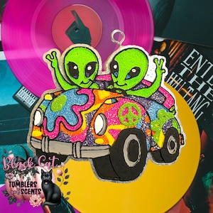 May include: A colorful air freshener featuring two green aliens in a psychedelic car with a peace sign. The air freshener is decorated with glitter and has a silver loop for hanging. The background includes a pink vinyl record and album art.
