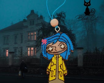 May include: A blue-haired, yellow-coated character with a stitched-up face, a pink flower, and white star-shaped eyes. The character is a handmade charm with a blue string and a wooden bead.