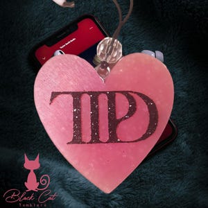 May include: A pink heart-shaped pendant with the letters "TIP" in dark glitter. The pendant hangs from a clear bead and cord. A smartphone displaying "Taylor Swift Radio" is visible in the background.