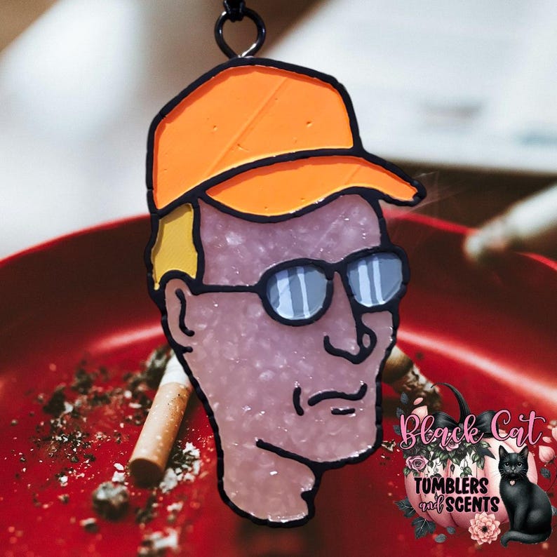 May include: A stained glass style charm of a cartoon man wearing an orange baseball cap and sunglasses. The charm is hanging from a black chain.