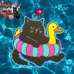 May include: A black cat-shaped air freshener with green eyes, lounging in a colorful inflatable ring with a yellow duck head. The ring has pink and teal stripes. A pink bead hangs from a black cord.