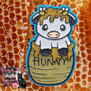 May include: A glittery, cartoon-style cow figure with black and white coloring, covered in gold honey drips, sitting inside a tan honey pot labeled HUNNY in black letters. The design features a bright blue border and a textured, sparkling finish.