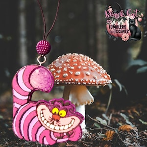 May include: A pink and purple Cheshire Cat-shaped ornament with a smiling face and striped body. The ornament hangs from a dark purple string with a matching beaded accent. A red mushroom with white spots is in the background. The logo "Black Cat Tumblers and Scents" is visible.