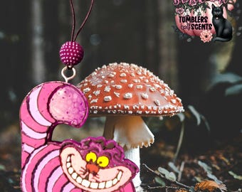 Op de afbeelding: Een roze en paarse Cheshire Cat-vormige ornament met een lachend gezicht en gestreept lichaam. Het ornament hangt aan een donkerpaarse draad met een bijpassend kralenaccent. Een rode paddenstoel met witte stippen staat op de achtergrond. Het logo "Black Cat Tumblers and Scents" is zichtbaar.
