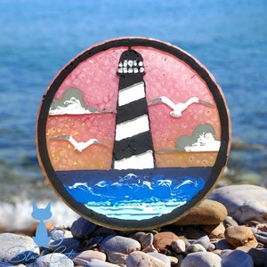 May include: A round, painted resin coaster with a lighthouse, seagulls, and ocean waves. The lighthouse is black and white with a red sky behind it. The coaster is on a bed of small rocks.
