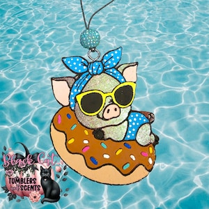 May include: A pig wearing sunglasses and a blue polka dot headband and swimsuit floats on a donut-shaped floatie. The pig is an air freshener with a black string and a glittery blue bead for hanging. The image has the text "Black Cat Tumblers and Scents".