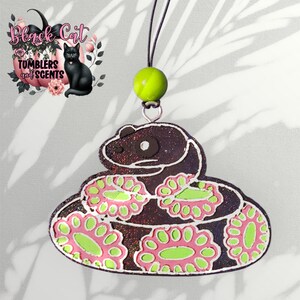 May include: A glittery, snake-shaped air freshener with pink and green floral accents. The air freshener is brown with a silver outline and a green bead at the top. The image also includes a logo with a black cat and the text "Black Cat Tumblers and Scents."