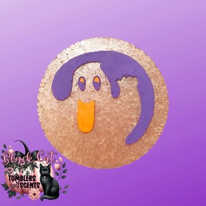 May include: A round, textured, gold-colored item with a purple design resembling a cartoon face. The face has two orange eyes, a purple head, and an orange tongue. The image also includes a logo with a black cat and the text "Black Cat Tumblers and Scents."