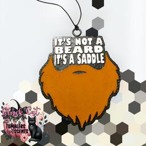 May include: A novelty air freshener shaped like a beard, with the text "IT'S NOT A BEARD IT'S A SADDLE". The beard is orange with a black outline. The air freshener is hanging from a black string. The logo "Black Cat Tumblers and Scents" is in the lower left.