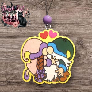 May include: A yellow heart-shaped ornament with two gnomes hugging. The female gnome is wearing a pink dress and a purple hat. The male gnome is wearing a green hat and a blue shirt. The ornament has two pink hearts above the gnomes.