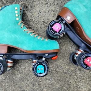 May include: Teal suede roller skates with black wheels and brown accents. The wheels have Pac-Man ghost designs in pink, yellow, and blue.