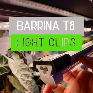 May include: A close-up of a black and white light clip for a T8 fluorescent light fixture. The clip is designed to hold the light fixture securely in place. The text "BARRINA T8 LIGHT CLIPS" is visible in the background.