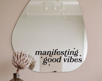 May include: An irregular, organic-shaped wall mirror featuring black vinyl lettering that reads "manifesting good vibes" accompanied by small star icons. The mirror reflects the surrounding room and is mounted on a plain, neutral-colored wall.