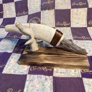 Peut inclure: Un couteau décoratif doté d'une lame en pierre grise fixée à un manche en bois de cerf clair avec un enroulement de cordon marron. Le couteau repose sur un petit support en bois de cerf clair, monté sur une base rectangulaire en bois brûlé.