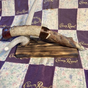 Puede incluir: Una pieza decorativa que presenta una base de asta de color claro que sostiene una hoja de piedra horizontal y puntiaguda. La hoja está asegurada al asta con una envoltura de color marrón oscuro. Todo el conjunto descansa sobre un bloque de madera quemada.