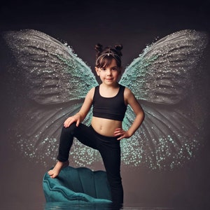 Puede incluir: Una ni&ntilde;a peque&ntilde;a con un top negro y leggings negros se para en una silla azul turquesa con grandes alas de mariposa azul claro transl&uacute;cidas detr&aacute;s de ella.