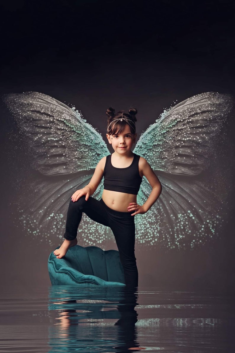 Puede incluir: Una ni&ntilde;a peque&ntilde;a con un top negro y leggings negros se para en una silla azul turquesa con grandes alas de mariposa azul claro transl&uacute;cidas detr&aacute;s de ella.