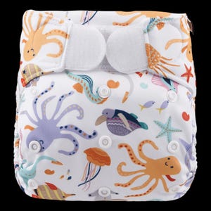 May include: A white cloth nappy with a colourful ocean-themed print featuring octopuses, seahorses, jellyfish, and starfish. The nappy has adjustable hook and loop fastenings.