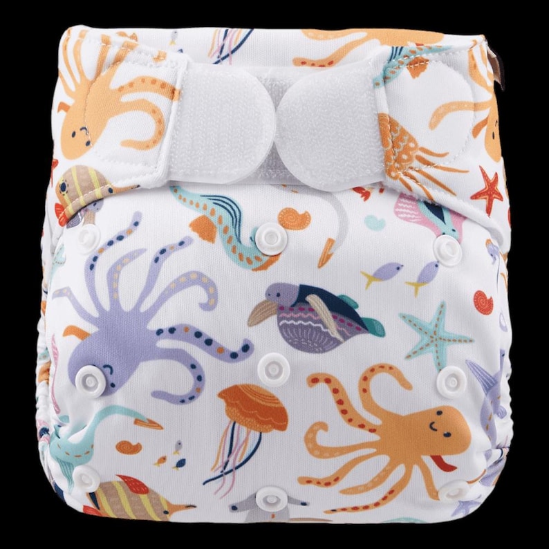 May include: A white cloth nappy with a colourful ocean-themed print featuring octopuses, seahorses, jellyfish, and starfish. The nappy has adjustable hook and loop fastenings.