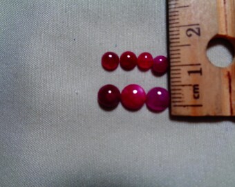 May include: Several polished, round gemstones in varying shades of red and pink. A wooden ruler with centimetre markings is placed beside the gemstones for scale. The gemstones are arranged on a light grey surface.